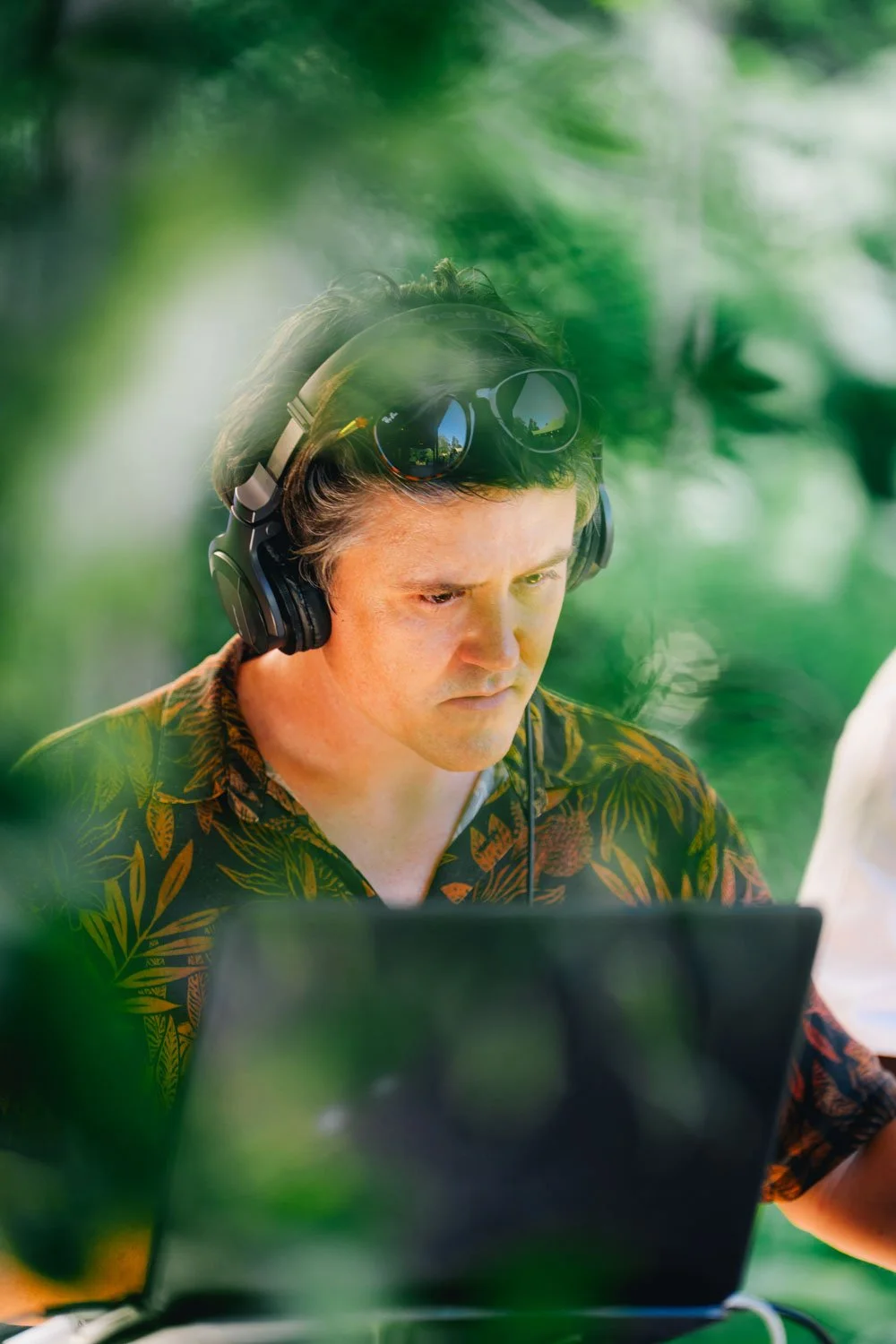 Man working outdoors with laptop, wearing headphones and sunglasses on his head, surrounded by greenery.