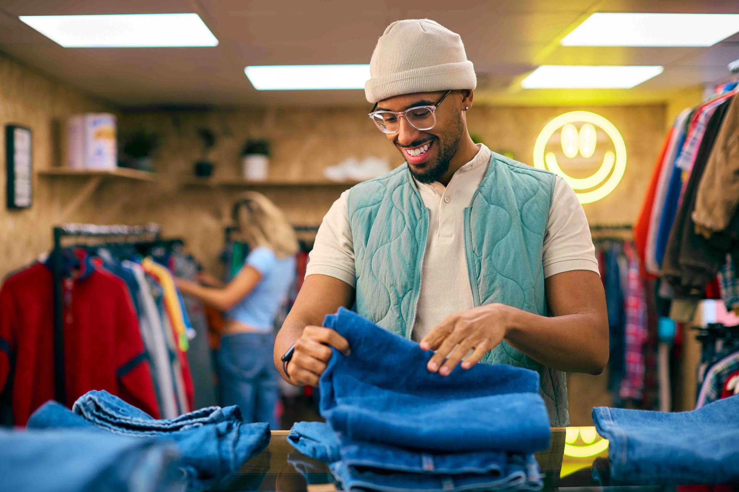 A young man with glasses and a beanie smiling and looking at clothes in a thrift store. There are racks of colorful clothing and a neon smiley face sign in the background.