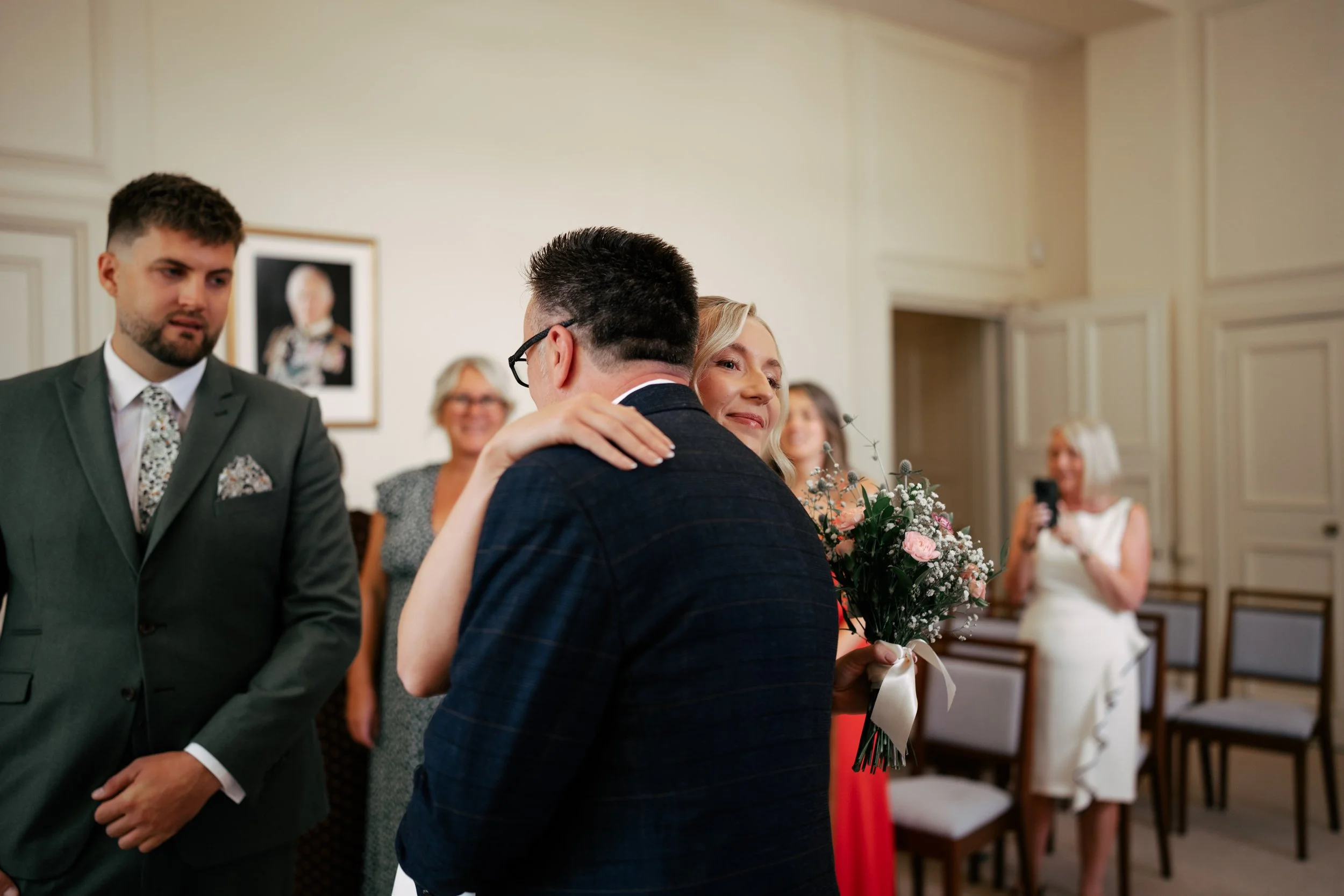 A woman in a red dress holding a bouquet hugging a man in a dark suit at a wedding reception.