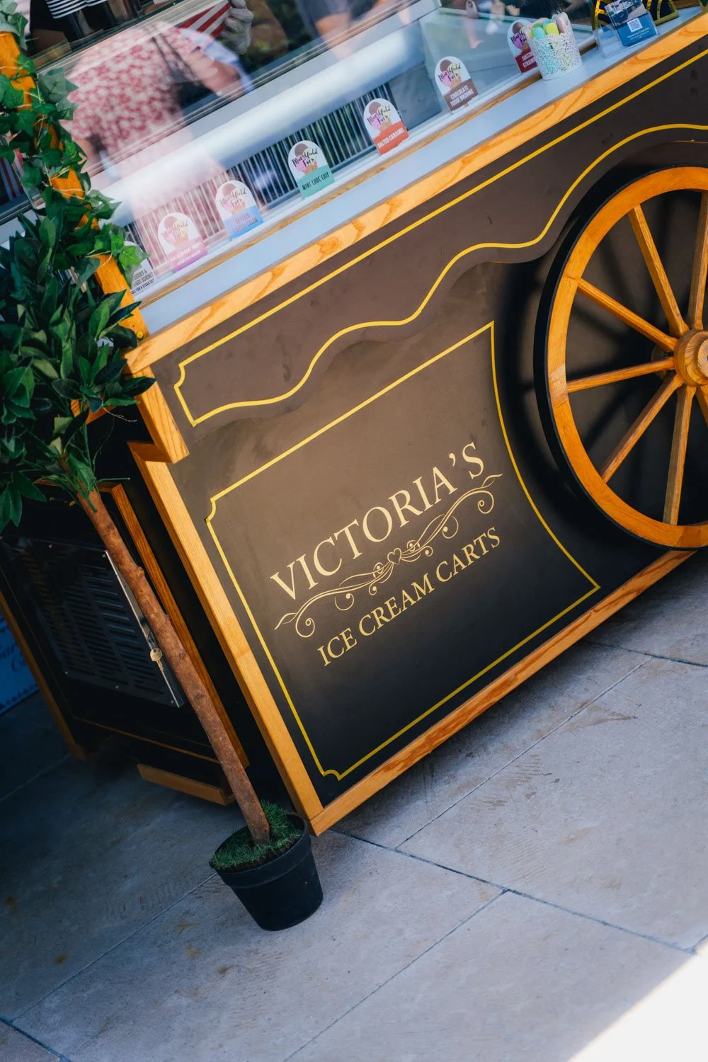 Sign on ice cream cart reading 'Victoria's Ice Cream Carts' with a decorative wheel and a potted plant next to it.