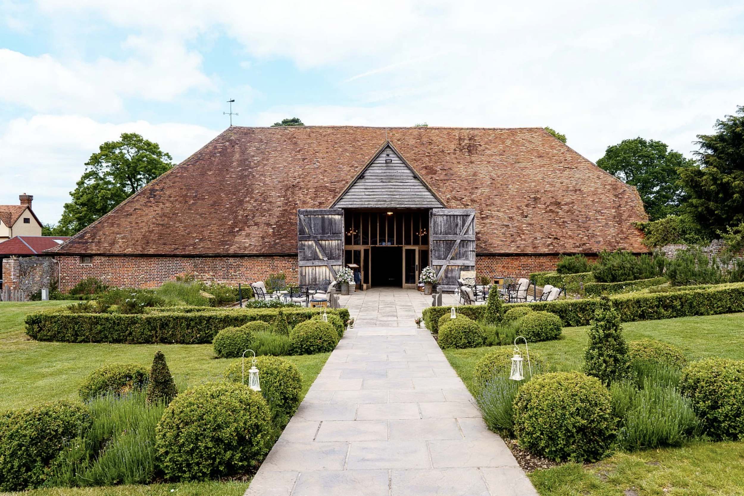 A rustic barn with large wooden doors open, situated in a well-manicured garden with symmetrical bushes and a stone pathway leading to the entrance, surrounded by lush green trees.