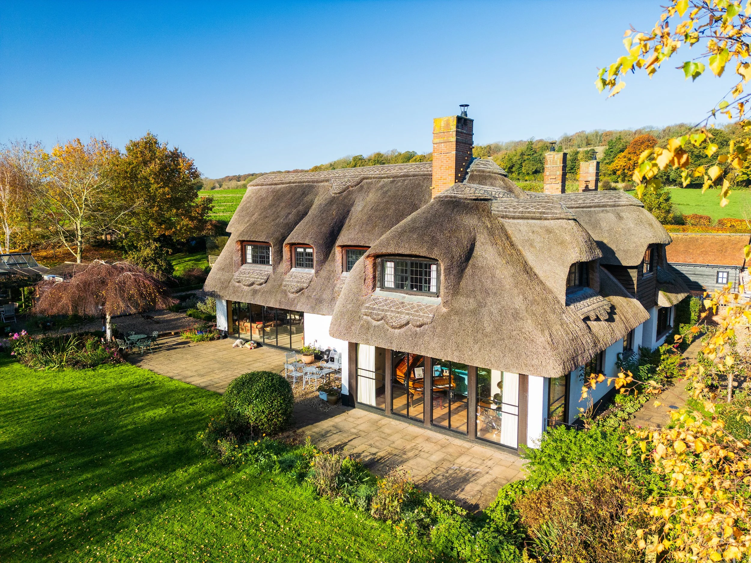 A house with a thatched roof, large glass windows, and surrounded by a well-maintained garden with trees and various plants.