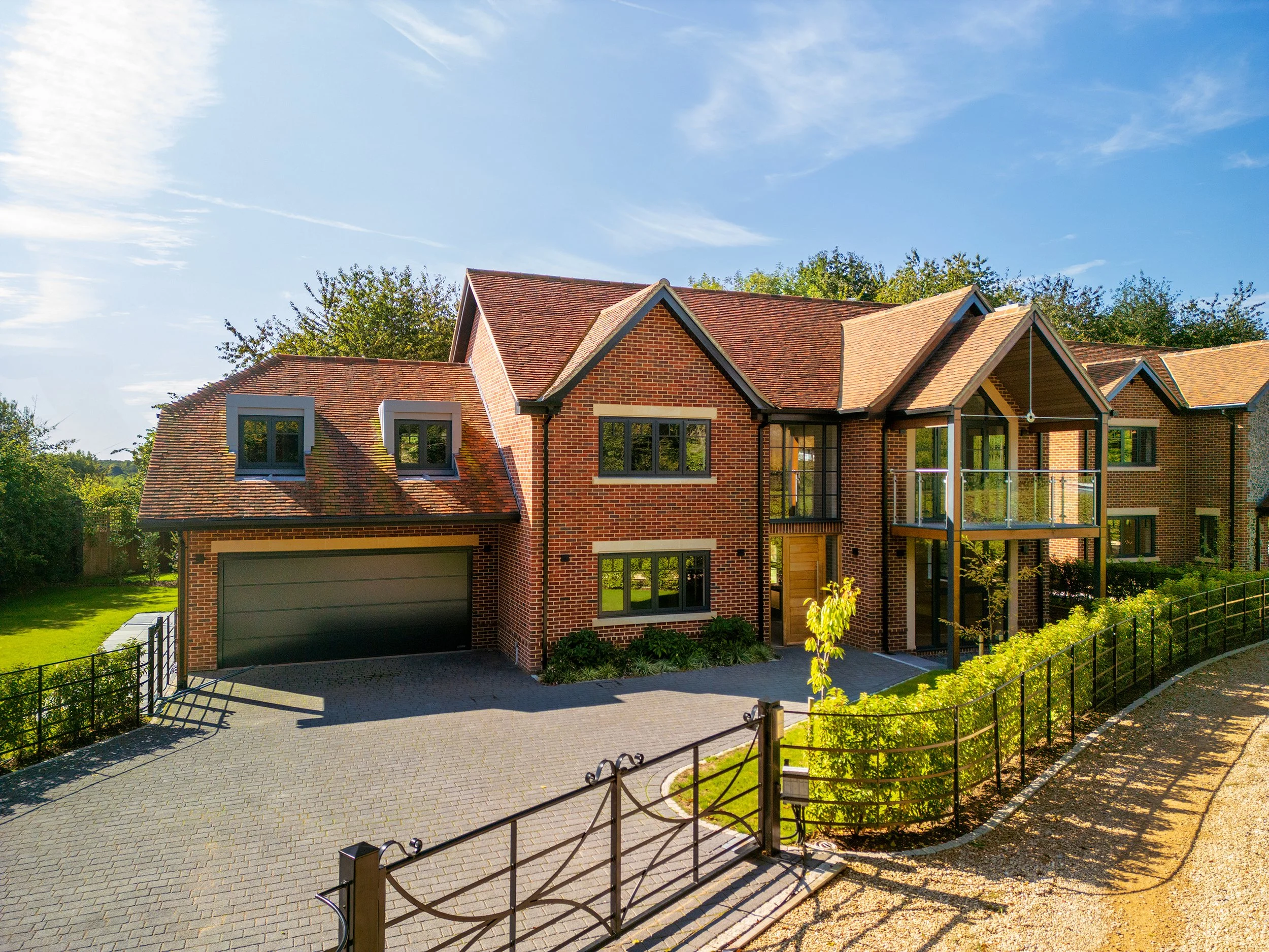 Modern brick house with multiple gabled roofs, large windows, and a balcony. Paved driveway with a black metal fence and green bushes.