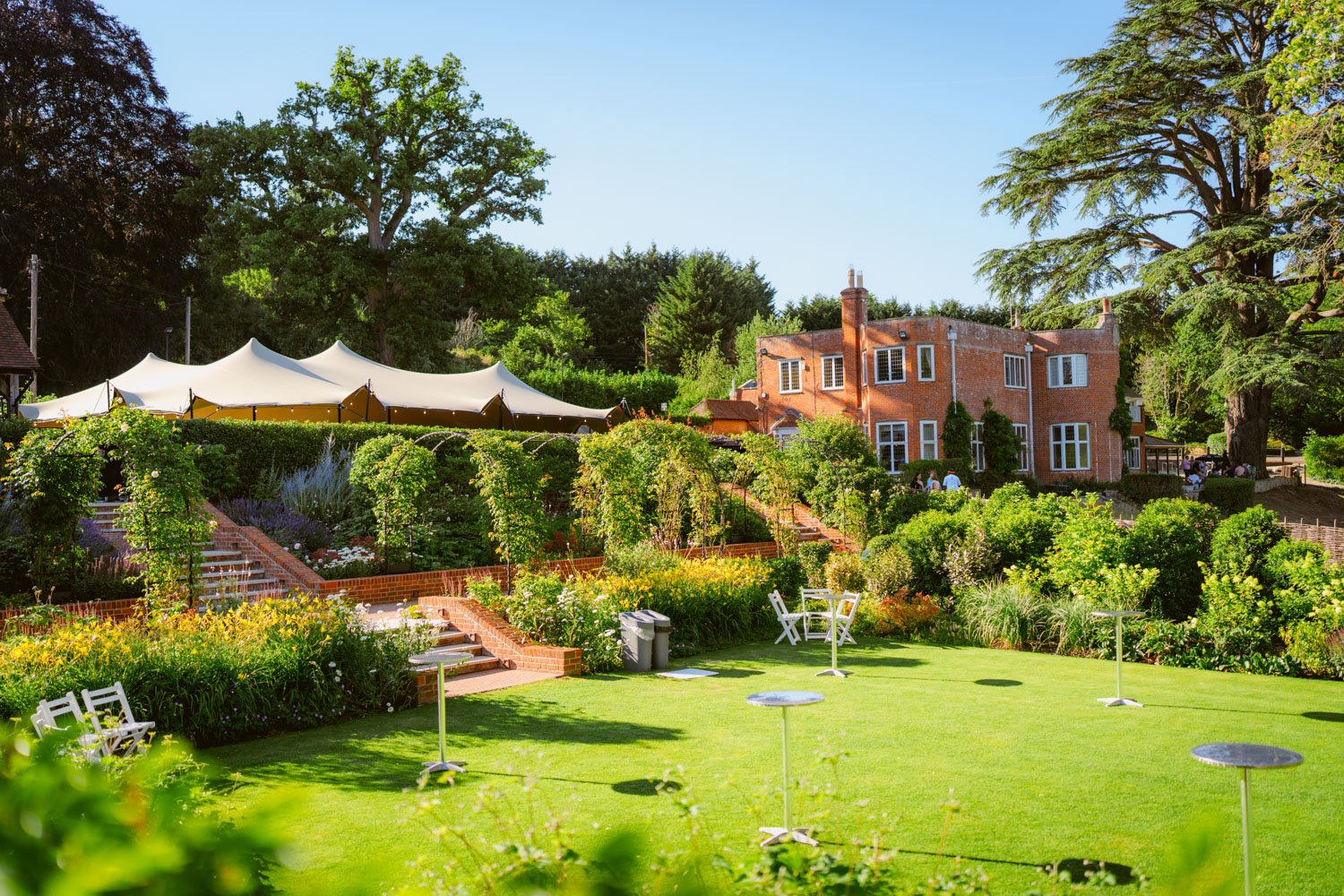 A lush garden with green grass, white benches, and small tables outdoors. There are steps and a variety of vibrant flowers and bushes leading up to a red brick building with large windows. A large white tent is partially visible on the left, shaded b