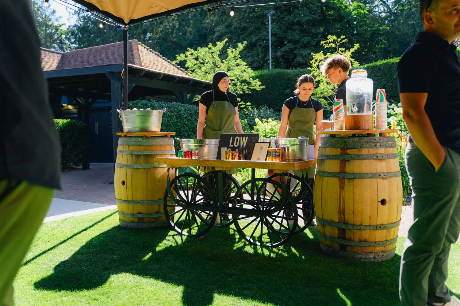 A beverage stand set up outdoors with wooden barrel tables, where two women wearing aprons are serving drinks. The stand has a sign that says 'LOW' and is surrounded by green trees and grass.