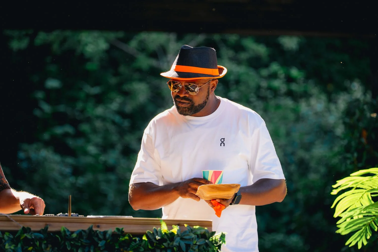 A man wearing a black fedora with an orange band, sunglasses, and a white t-shirt with a small logo, standing outdoors in a green environment, holding a paper or card. There is lush greenery around him.