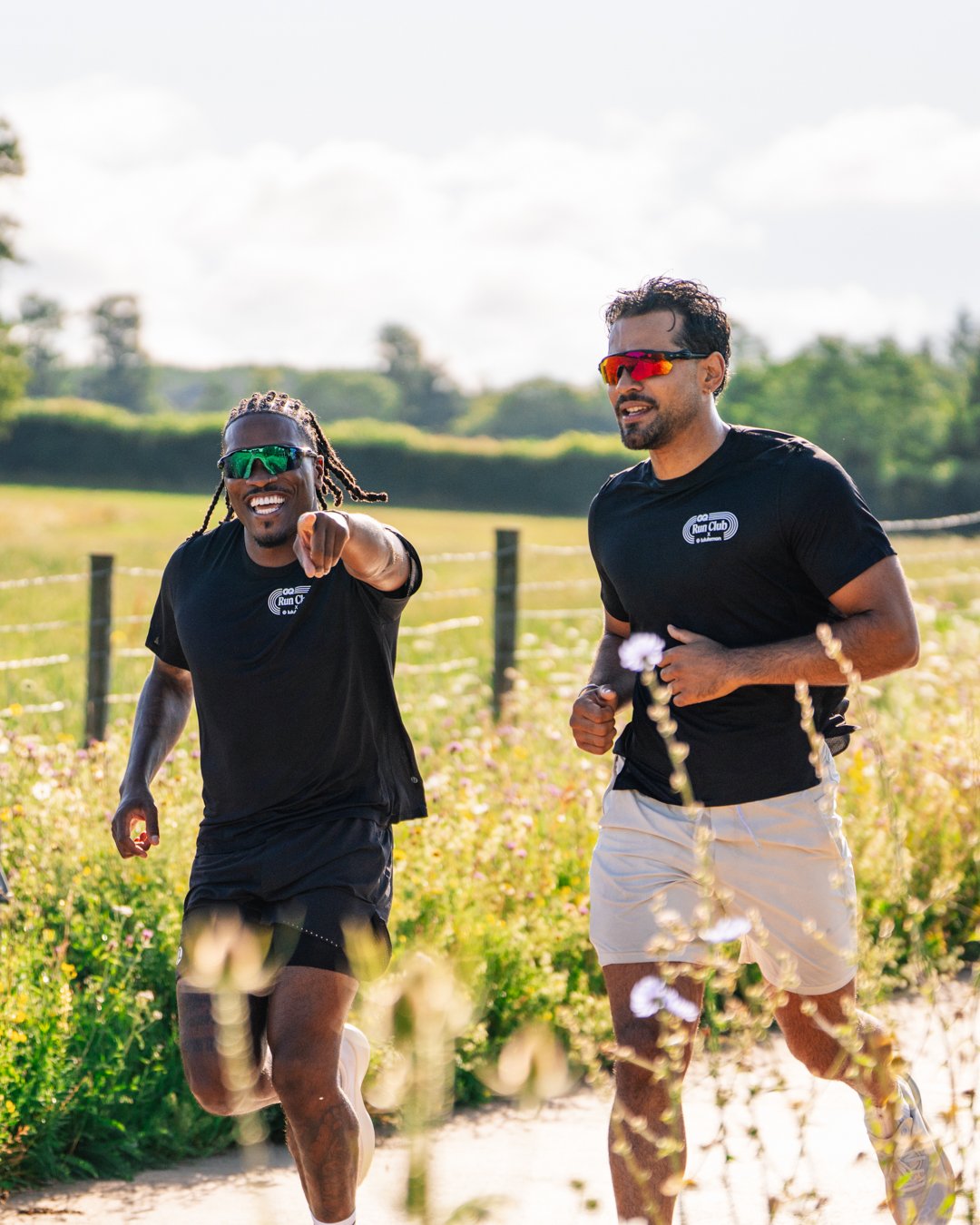 Two men running outdoors in a field during daytime, wearing black t-shirts and sunglasses, smiling and enjoying the activity.