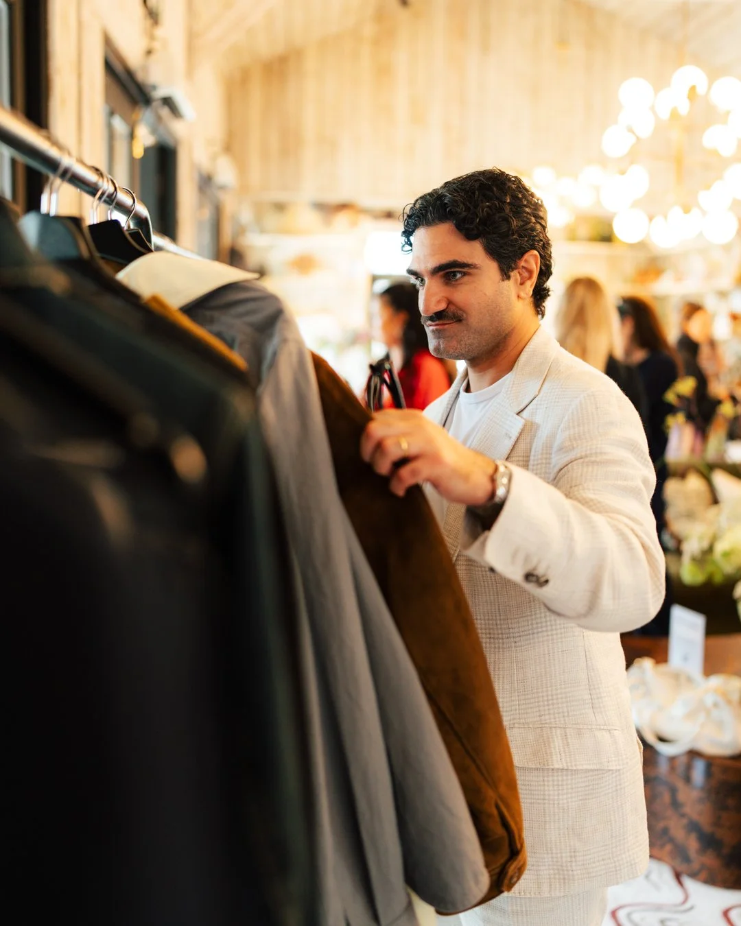 A man shopping for jackets at a clothing store, browsing through a rack filled with various jackets in a warmly lit shop.