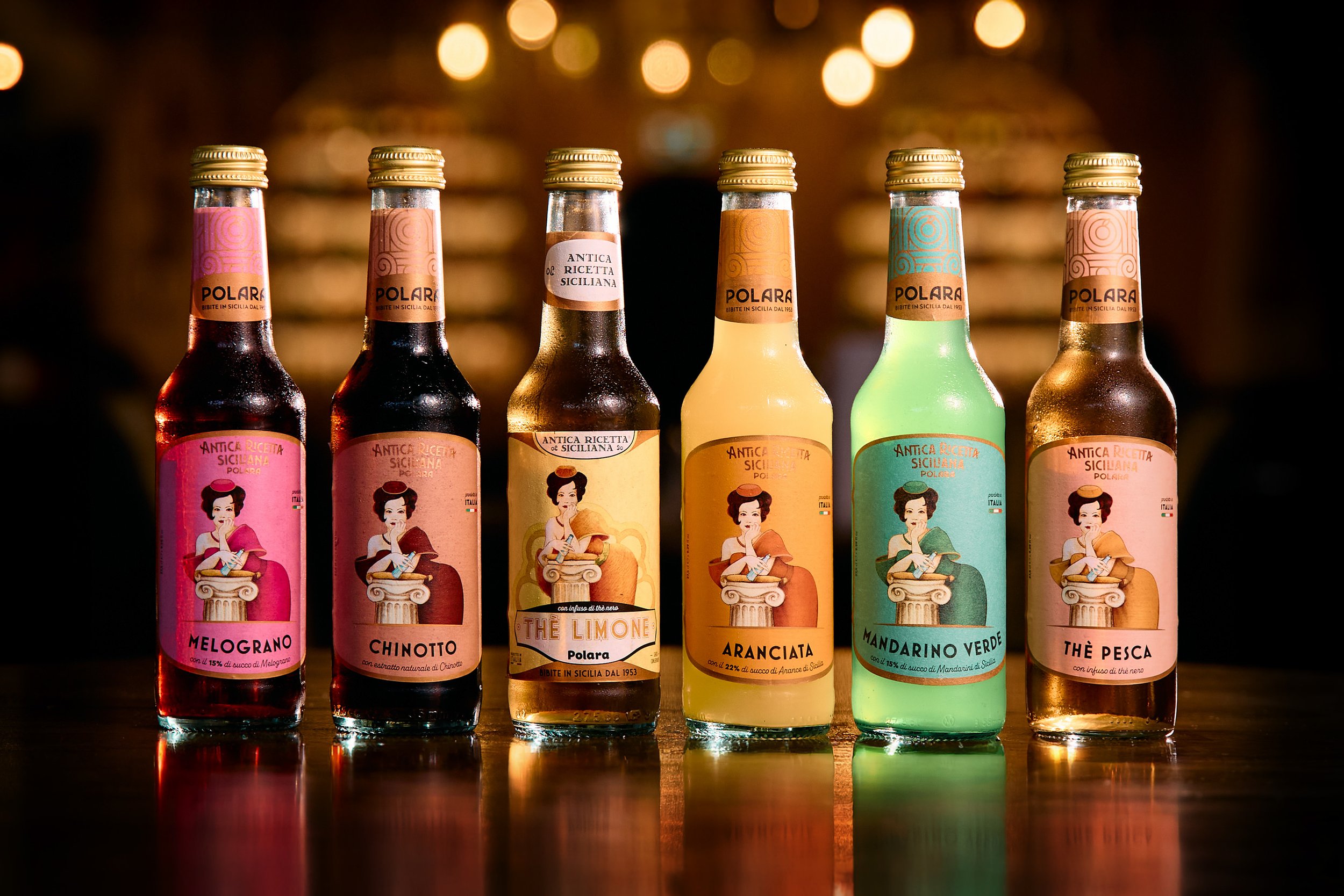 Six colorful bottles of Sicilian flavored liquors with illustrated labels, aligned on a wooden surface against a dark, bokeh background.
