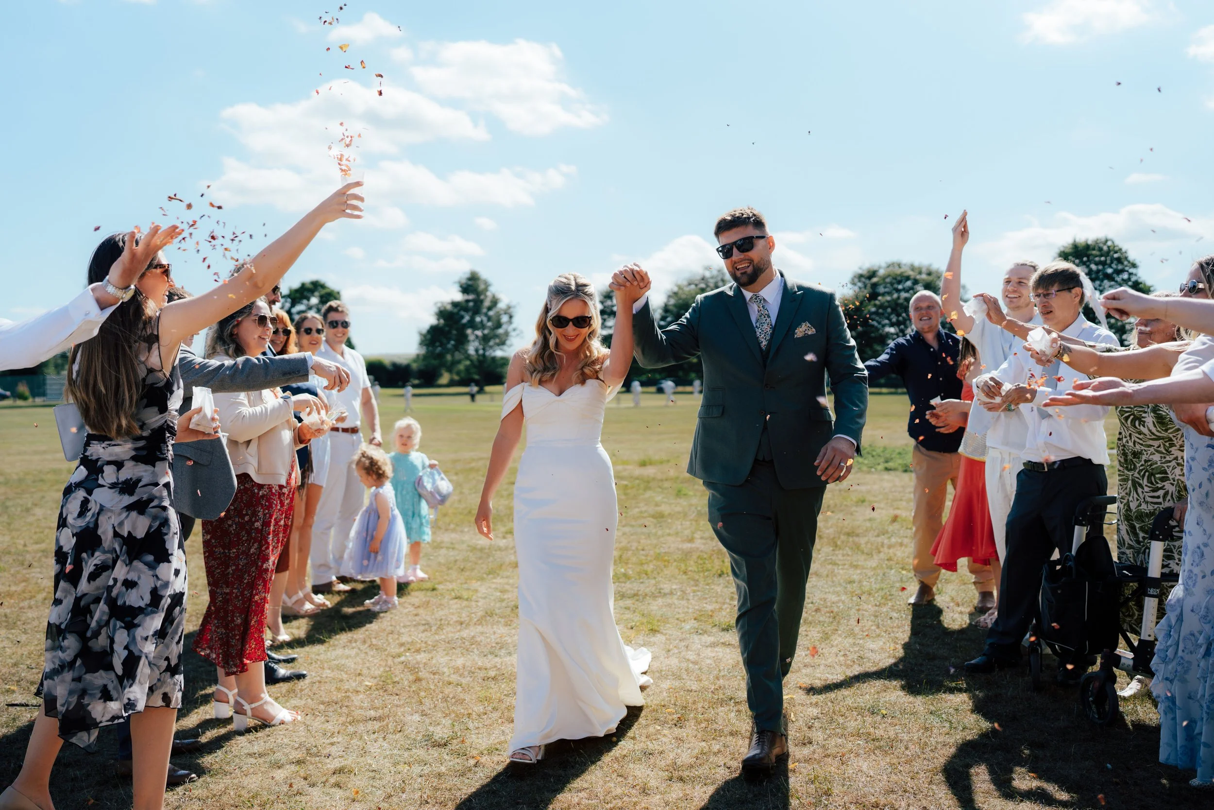 A bride and groom celebrating with guests outdoors in a sunny, open field during their wedding reception, throwing confetti.