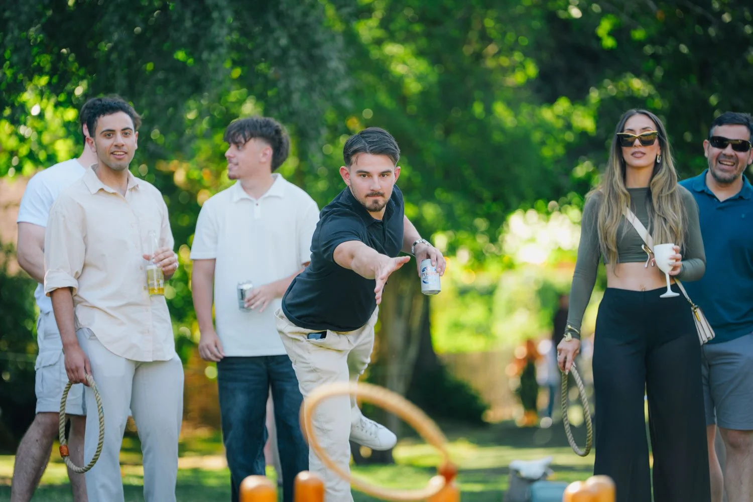 Group of people playing cornhole outdoors on a sunny day, with trees in the background. One man is throwing a bean bag, others are holding drinks and watching, some wearing sunglasses.