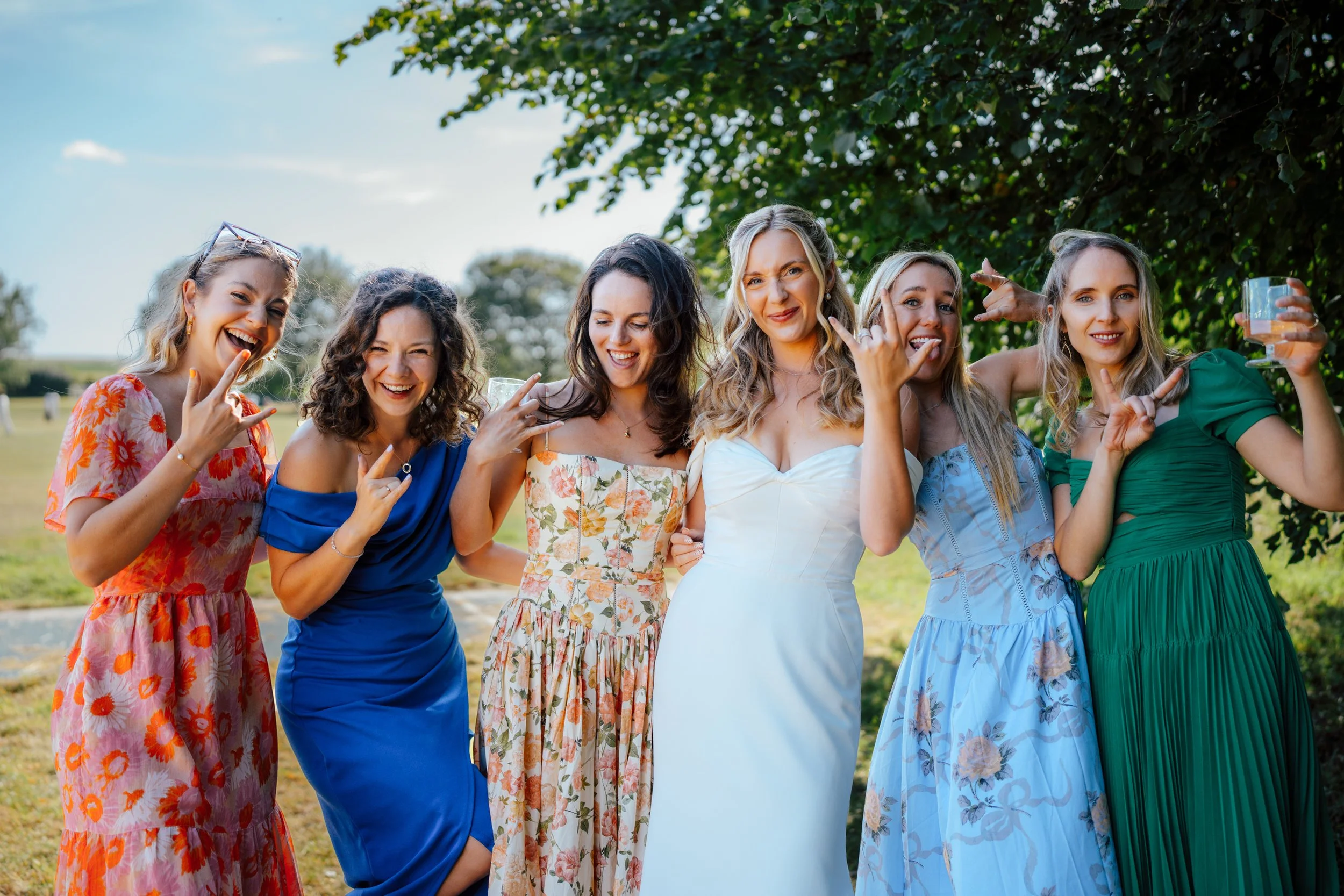 Seven women standing outdoors beneath a tree on a sunny day, smiling and making hand gestures, with some holding drinks.