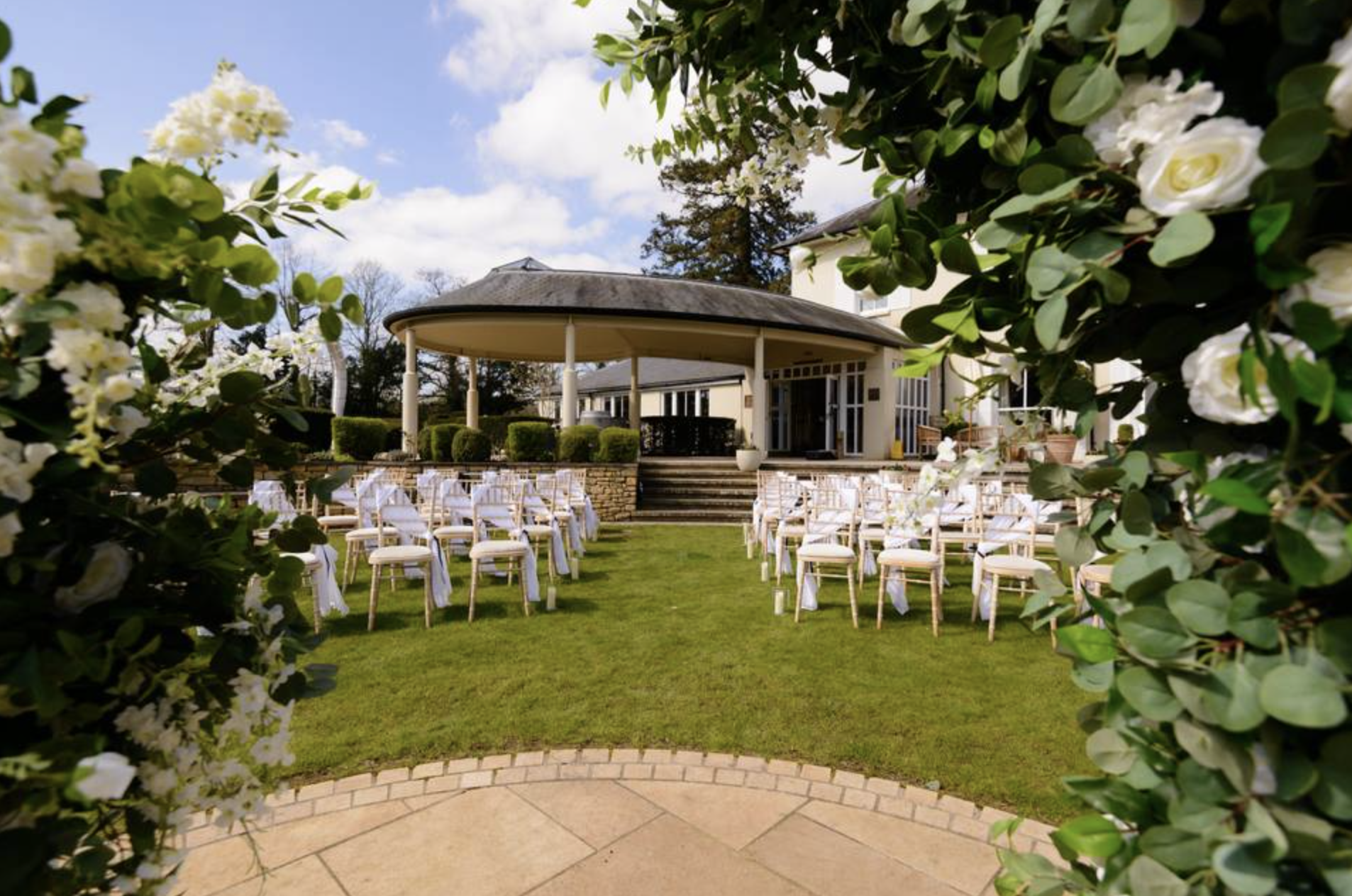 A wedding ceremony setup outdoors, with white chairs arranged on green grass facing a small stage with a white house and veranda in the background, framed by green foliage and white flowers.