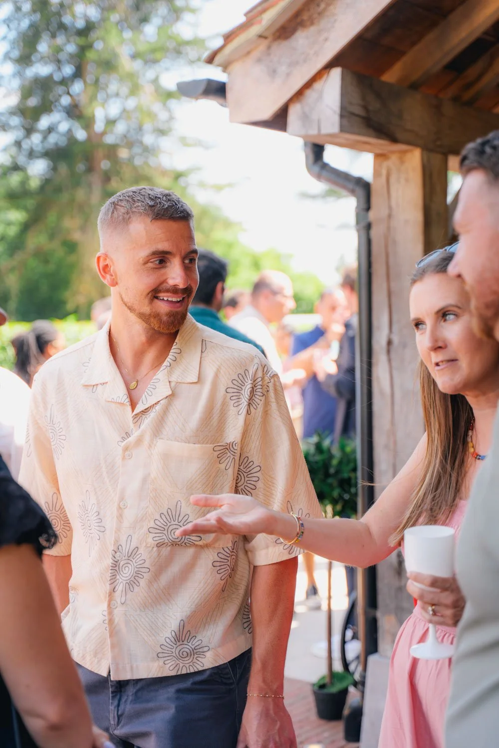 People socializing outdoors at a gathering, with a man in a beige patterned shirt smiling and talking to a woman holding a glass.