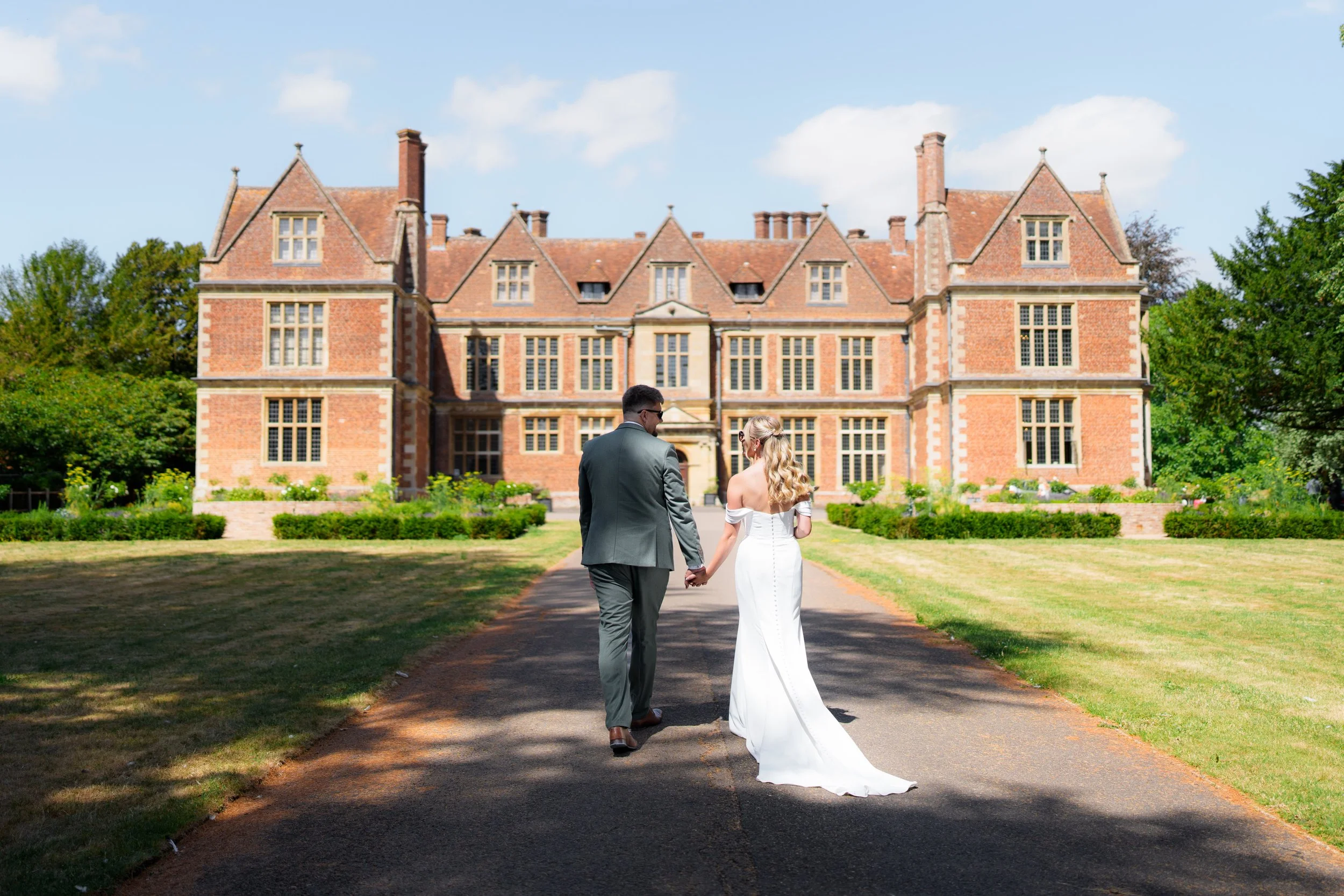A bride and groom holding hands and walking towards a large historic brick mansion or castle on a sunny day, surrounded by greenery.