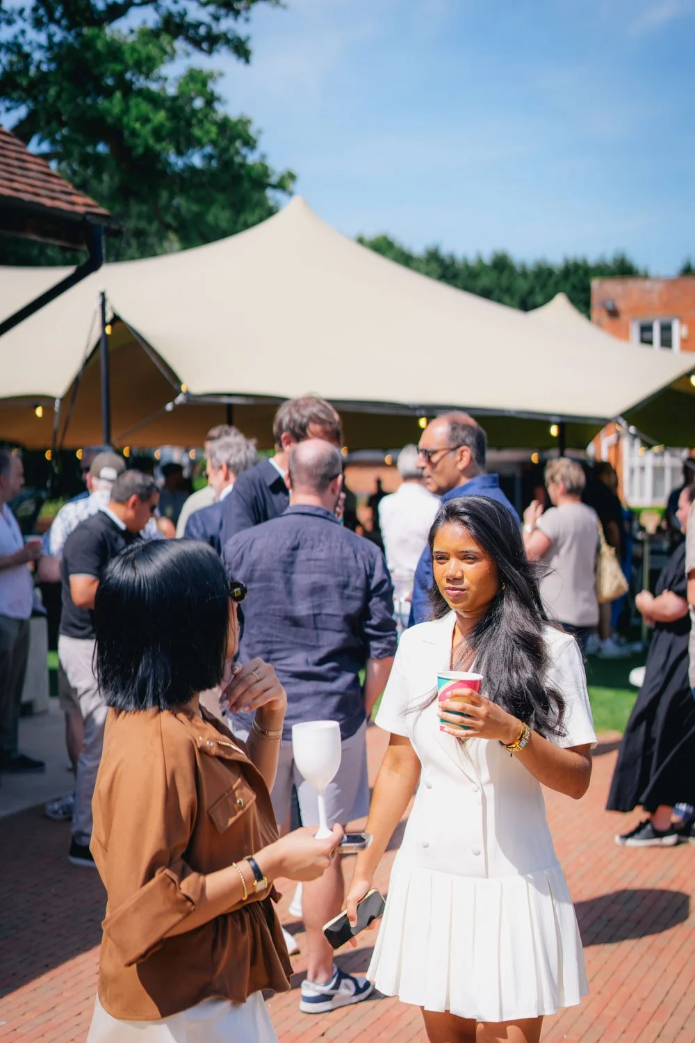 People gathered outdoors under a white canopy at an event on a sunny day, engaging in conversations, holding drinks, with some objects in hand.