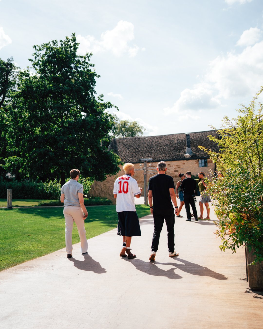 Group of people walking outside on a sunny day near a stone building with a tiled roof and trees, some dressed casually and others more formally.