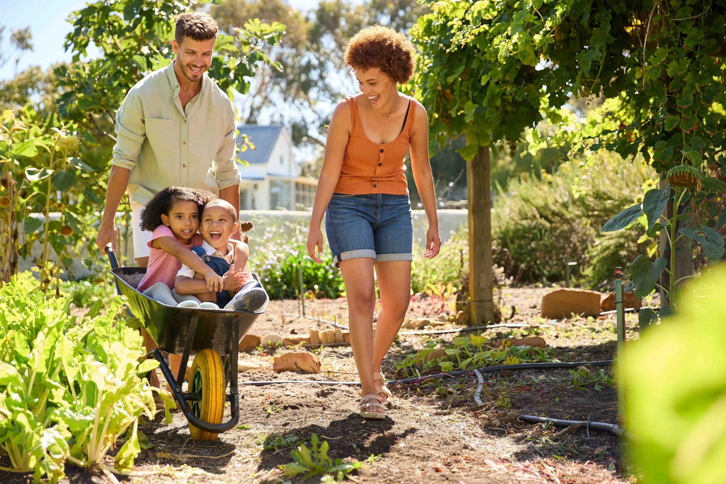 Family gardening in a lush backyard during daytime. A woman and two children, one in a wheelbarrow, are smiling and walking among green plants and vegetables, with trees and a house in the background.