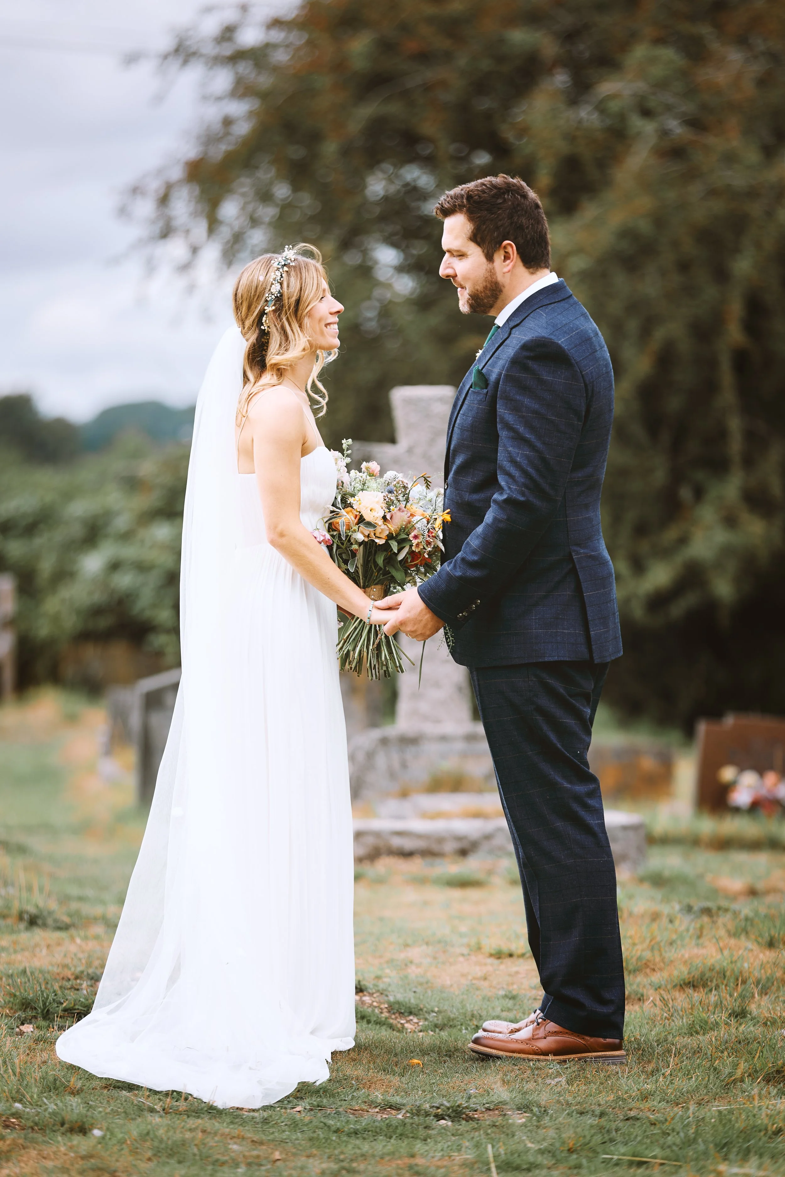 A bride and groom holding hands during their outdoor wedding, standing on grass with trees and gravestones in the background.