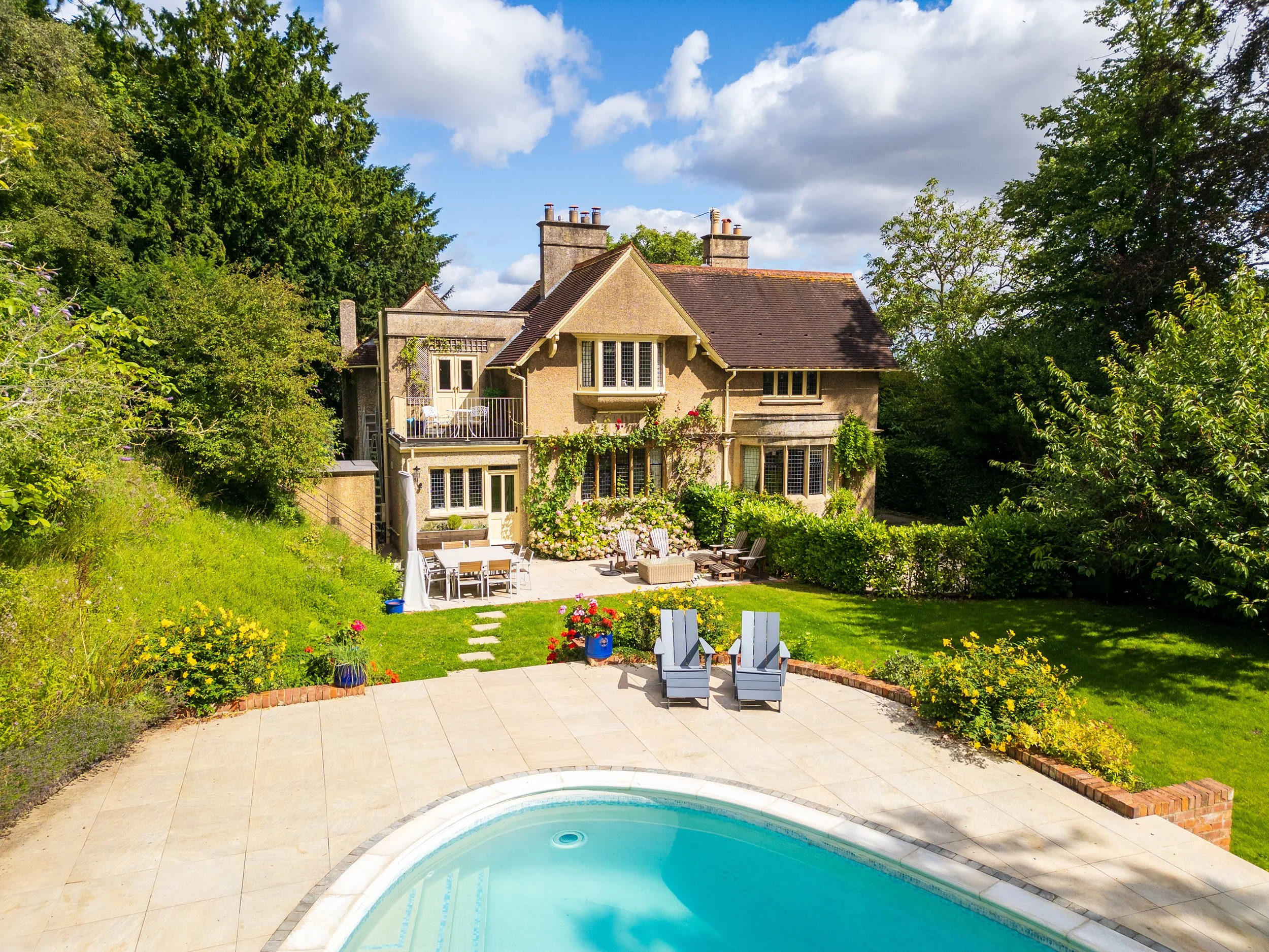 Backyard with a swimming pool, outdoor seating, and a large beige house surrounded by green trees and garden plants.