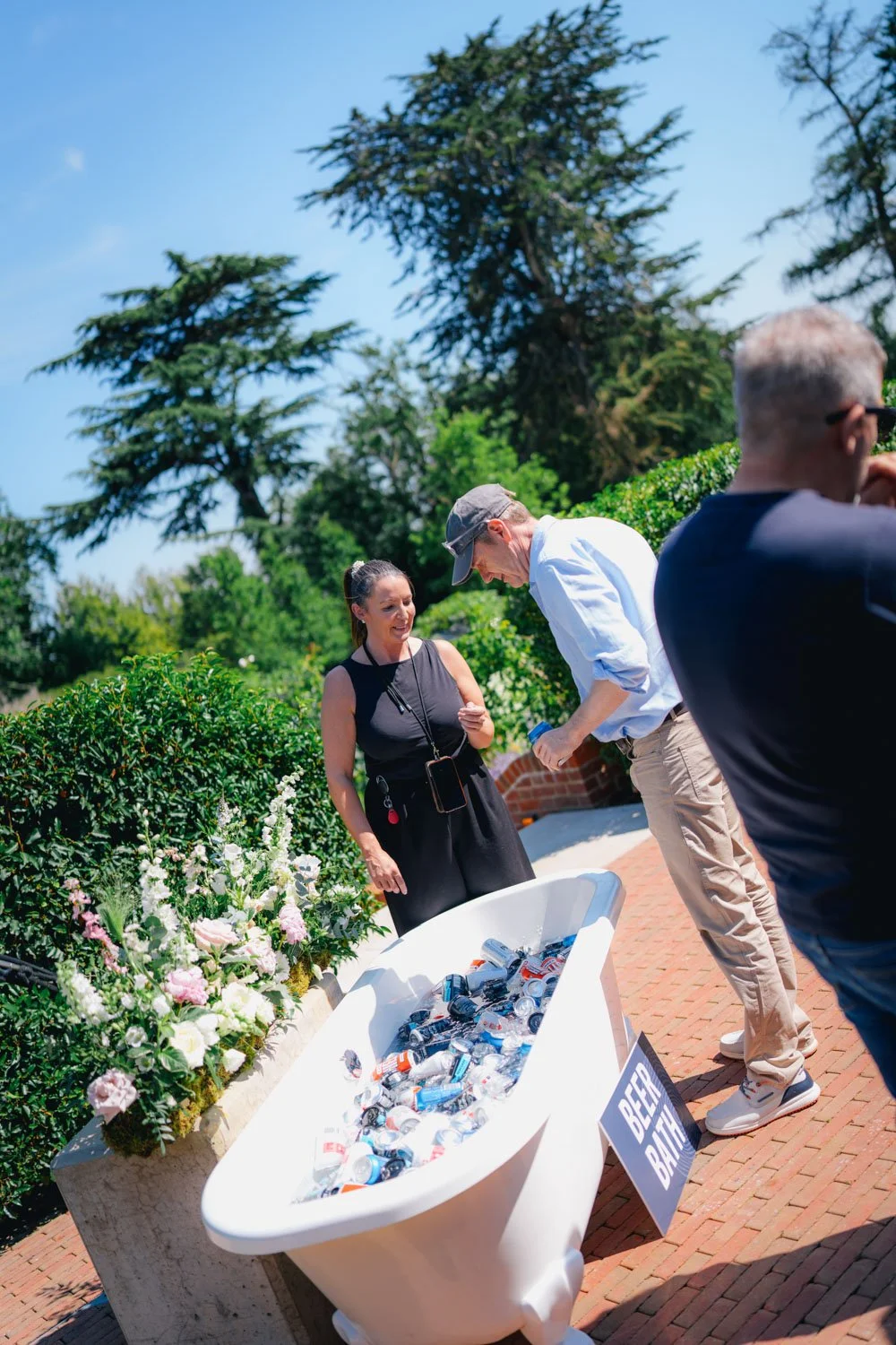 People participating in a beer bath event outdoors on a sunny day, with a bathtub filled with cans of beer, a sign reading "Beer Bath," and lush greenery in the background.