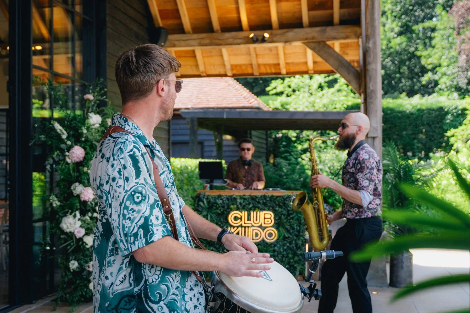 Three musicians performing outdoors at a venue named 'Club Kudo'. A man in Hawaiian shirt plays conga drums, a bald man with a beard plays saxophone, and in the background a DJ operates equipment, with lush greenery surrounding them.