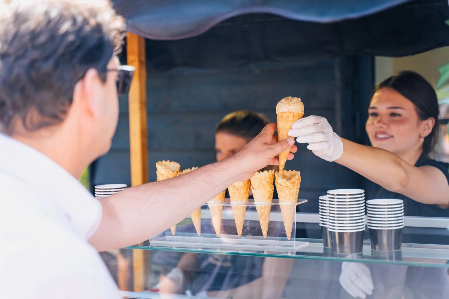 A customer at an ice cream stand is receiving a cone from a smiling female vendor behind the counter. The stand has multiple ice cream cones on display and stacks of paper cups.