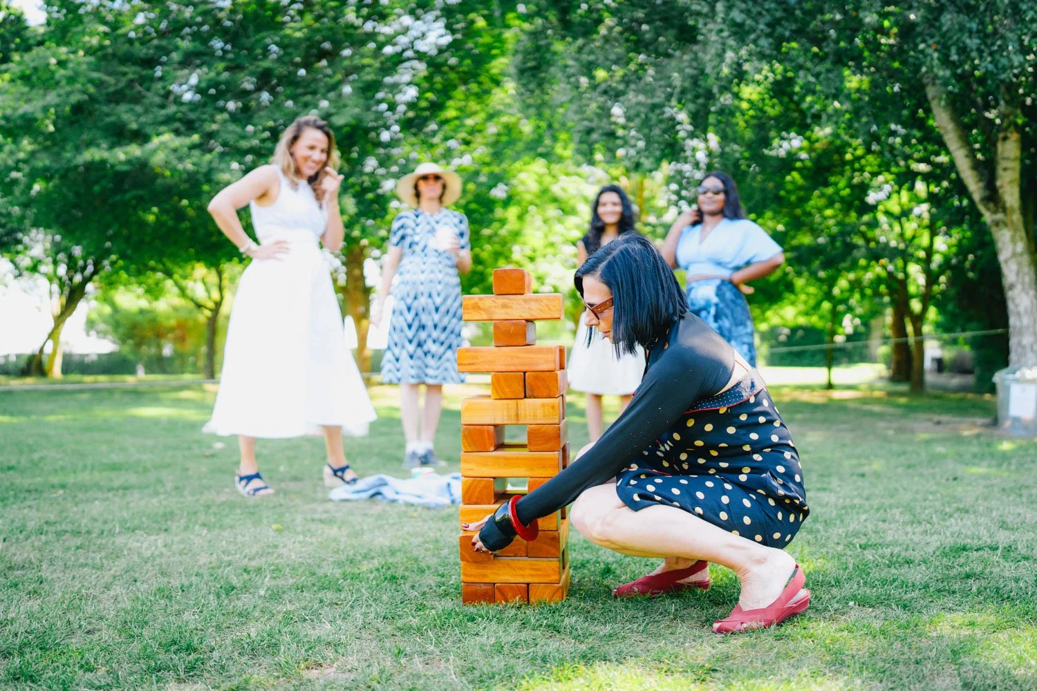 Women playing giant Jenga outdoors in a park, with green trees and sunlight in the background.