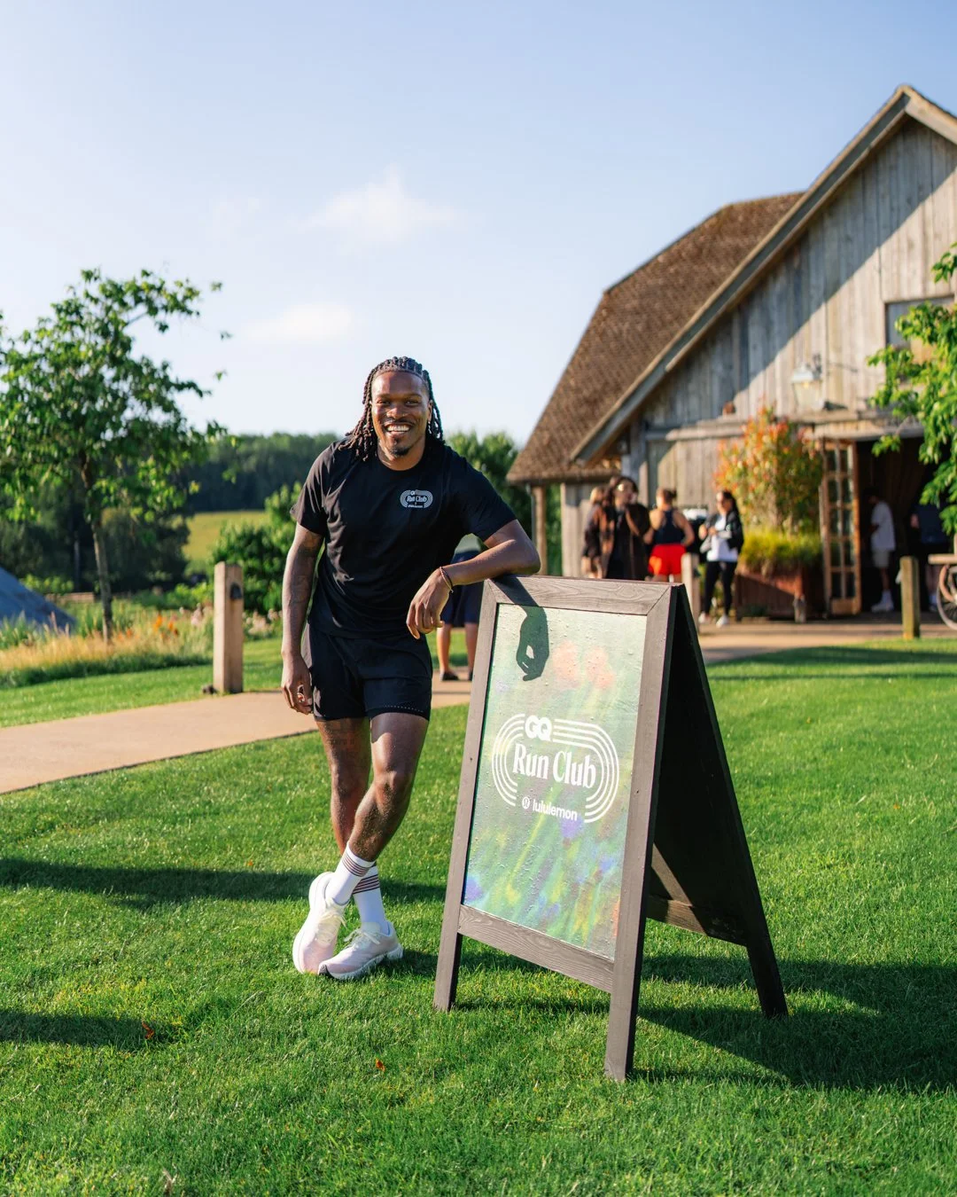 A man with dreadlocks wearing a black t-shirt and shorts smiling and leaning on a club sandwich board sign that reads "Run Club" outdoors near a rustic barn with a group of people in the background.