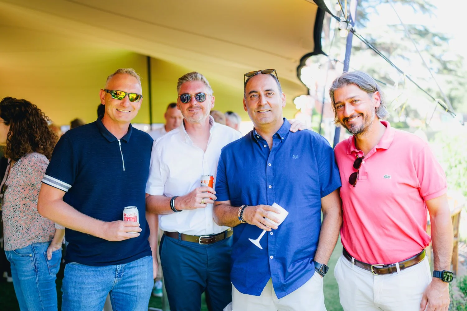 Group of four men smiling at an outdoor event, holding drinks, dressed casually in polo shirts and button-up shirts, some wearing sunglasses.
