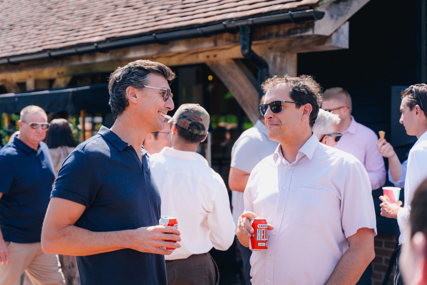 Two men wearing sunglasses and casual shirts are talking and holding canned beverages at an outdoor gathering, with other people in the background enjoying the event.