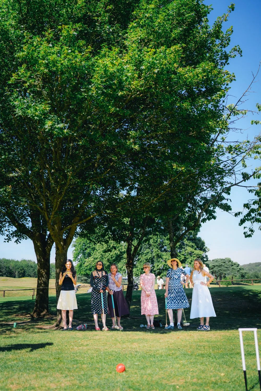 Group of women standing outdoors on a sunny day, laughing and smiling under a large tree, with a golf ball in the foreground.