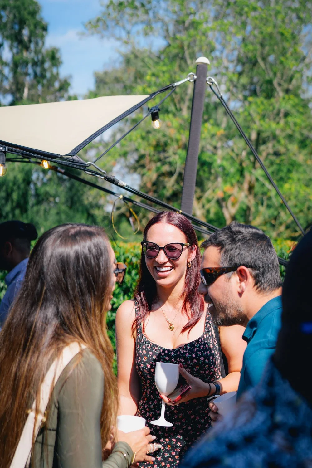 A group of people outdoors, smiling and talking at a social gathering with trees and a large canopy overhead.