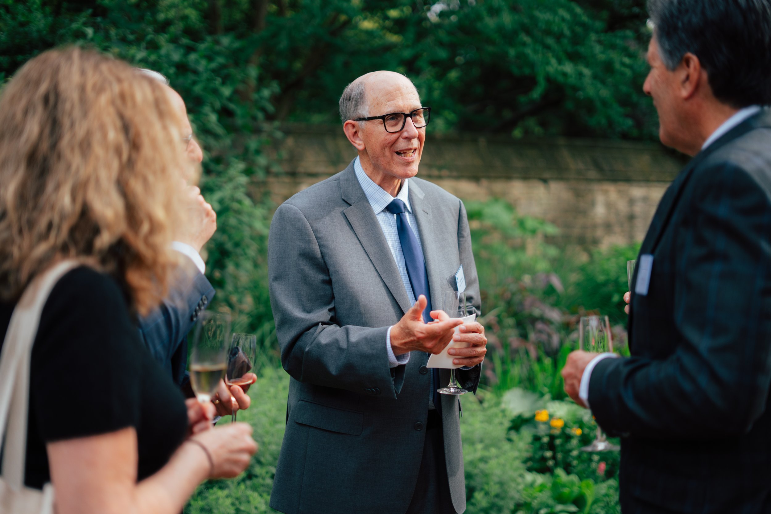 A group of people in formal attire having a conversation outdoors, with a man in glasses and a gray suit speaking, holding a glass of wine, during a social gathering in a park.