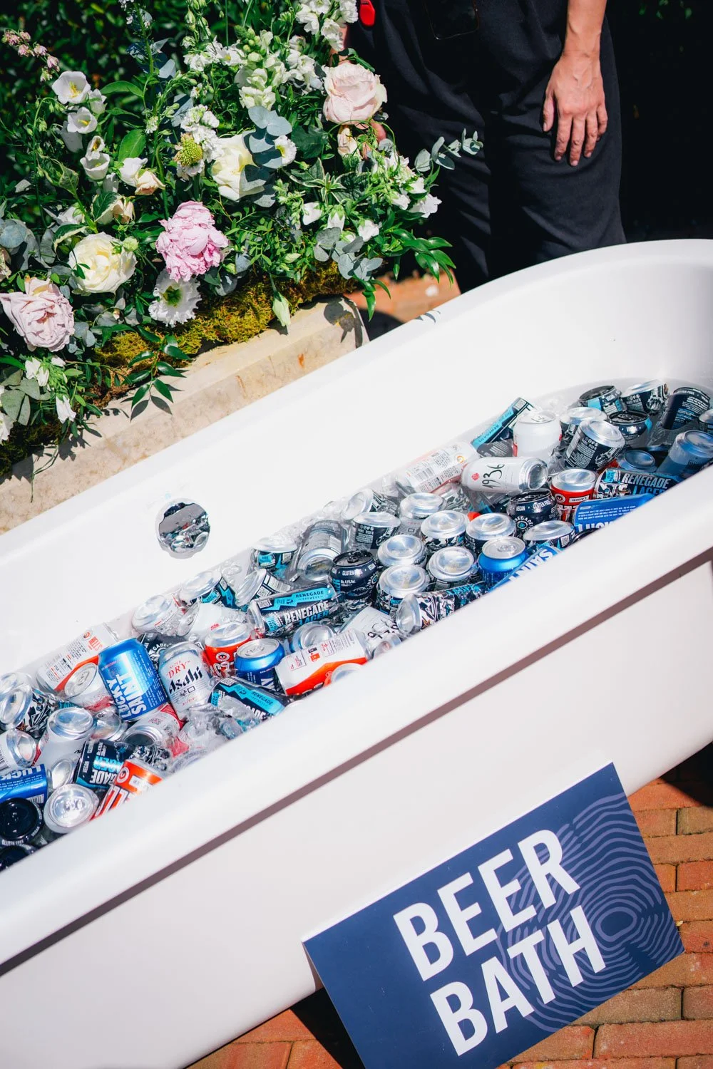 An outdoor beer bath filled with assorted cans and bottles of beer, next to a large floral arrangement and a sign that reads "Beer Bath."