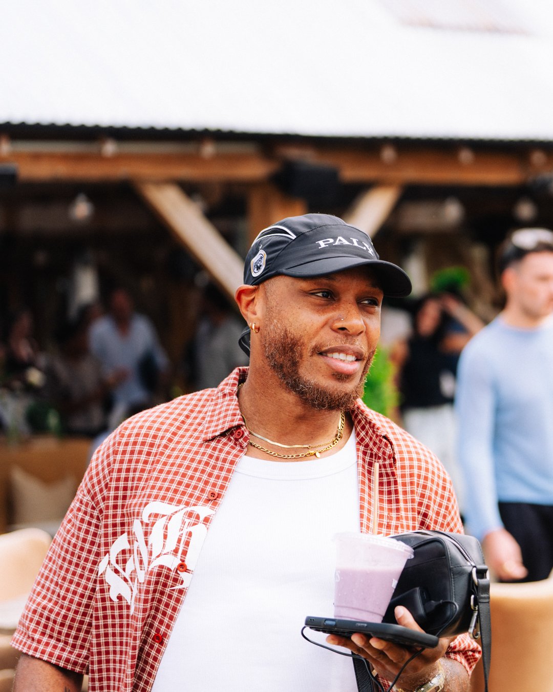 A man with a beard and earrings wearing a black cap, a red checkered shirt over a white t-shirt, holding a cup of pink smoothie, a phone, and a small black backpack at an outdoor event.