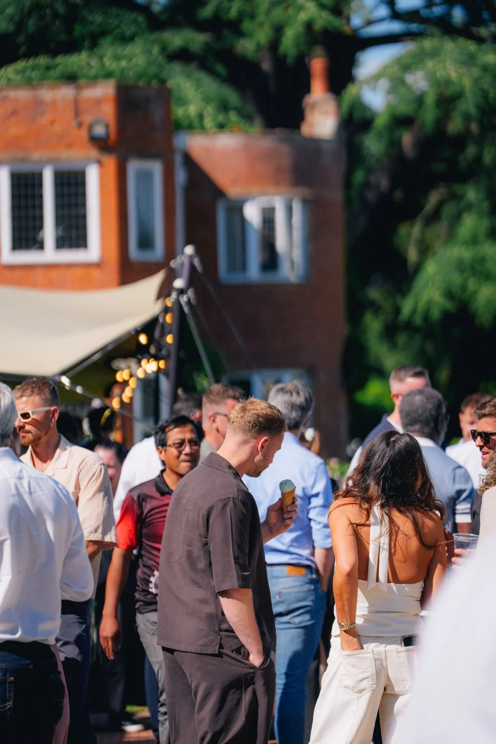A lively outdoor gathering with people socializing, wearing casual and semi-formal attire, some holding drinks or ice cream, with a tent decorated with string lights and a brick house with white window frames in the background on a sunny day.