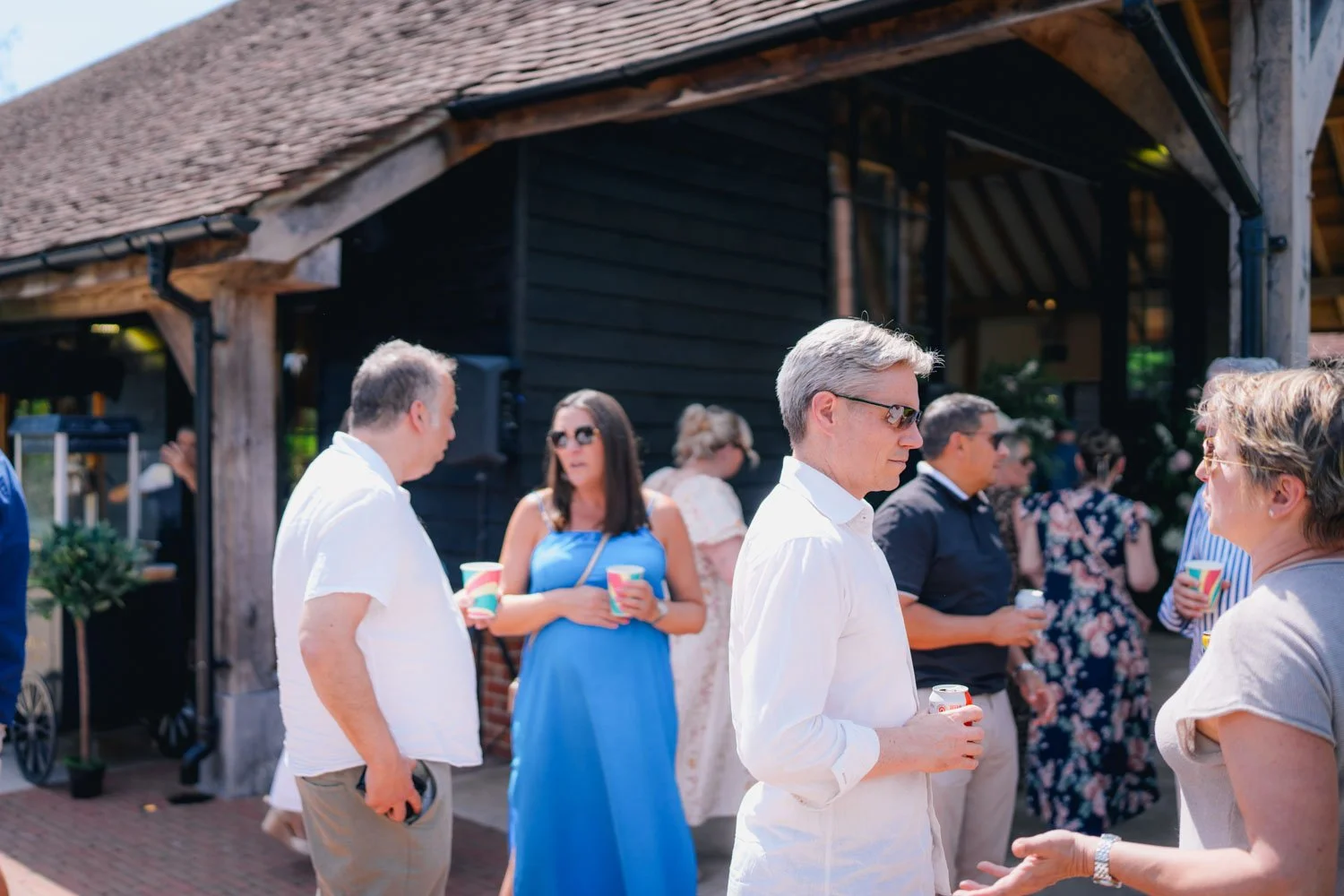 People socializing outdoors at a gathering, some holding cups and drinks, in front of a black wooden building with a tiled roof on a sunny day.