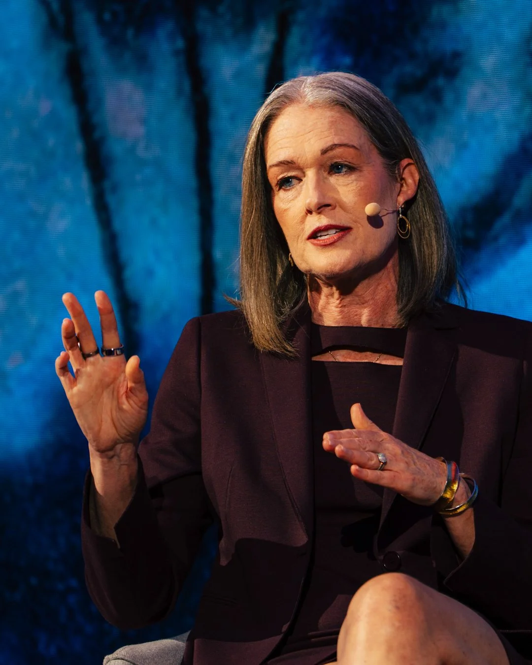 A woman with gray hair speaking at an event, wearing a dark blazer and using a microphone headset, with a blue and black textured background.