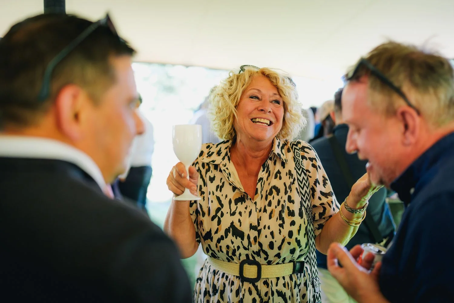A woman with curly blonde hair wearing a leopard print dress and holding a white wine glass, smiling and talking to two men at a social gathering.