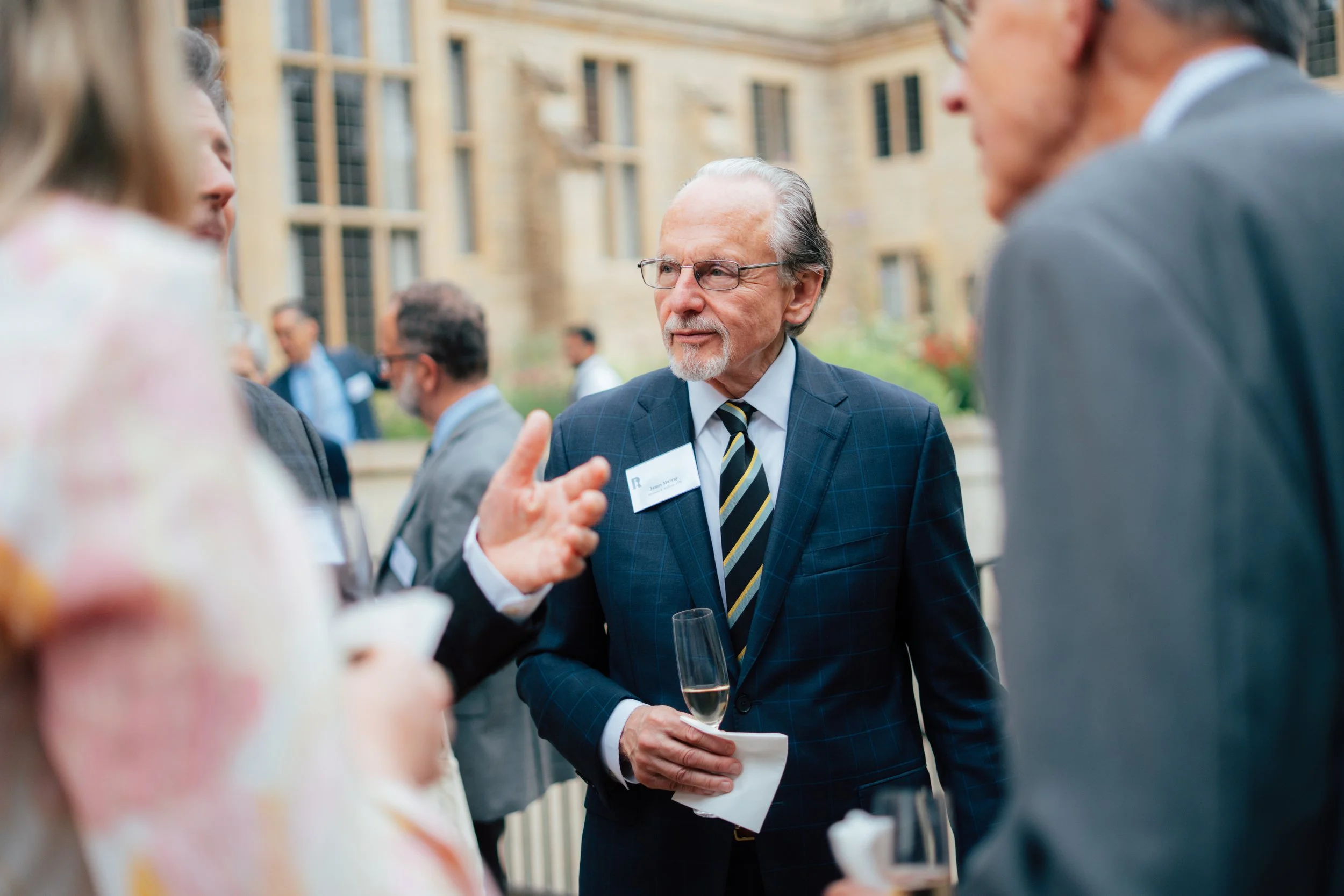 An elderly man in a suit and tie holding a glass of champagne, talking to people at an outdoor social event in front of a building with large windows.