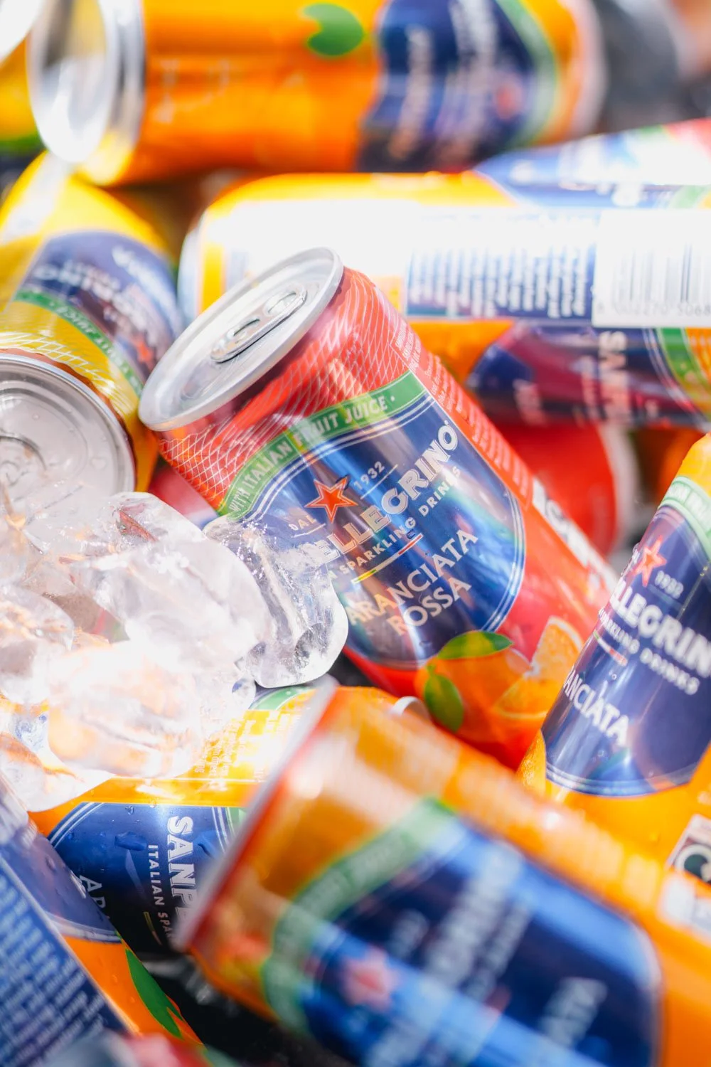 Close-up of colorful cans of Sanpelligrino sparkling drinks in a pile with ice cubes.