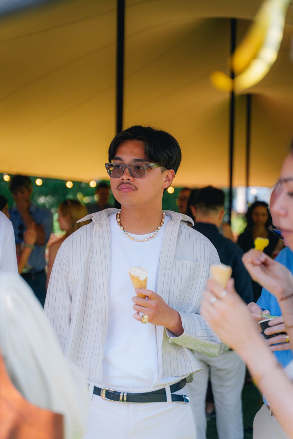 Young man with black hair, glasses, and a mustache wearing a white t-shirt, a light striped open shirt, and a pearl necklace, holding an ice cream cone at an outdoor gathering under a yellow tent with other people in the background.