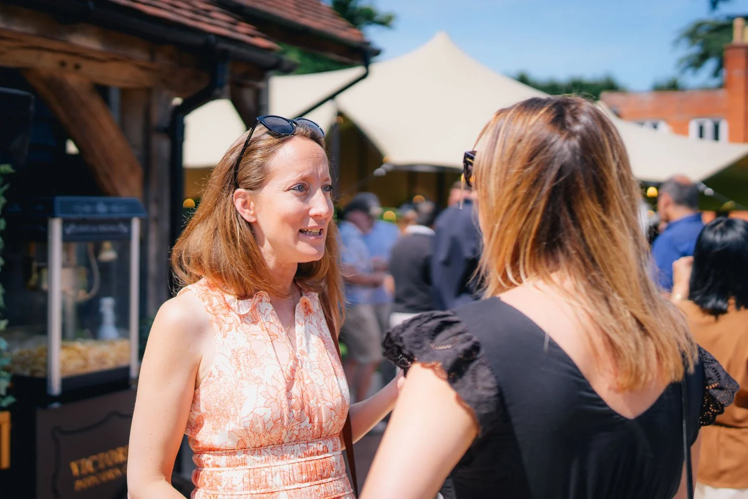 Two women conversing outdoors at a social event on a sunny day, with a crowd and large umbrellas in the background.
