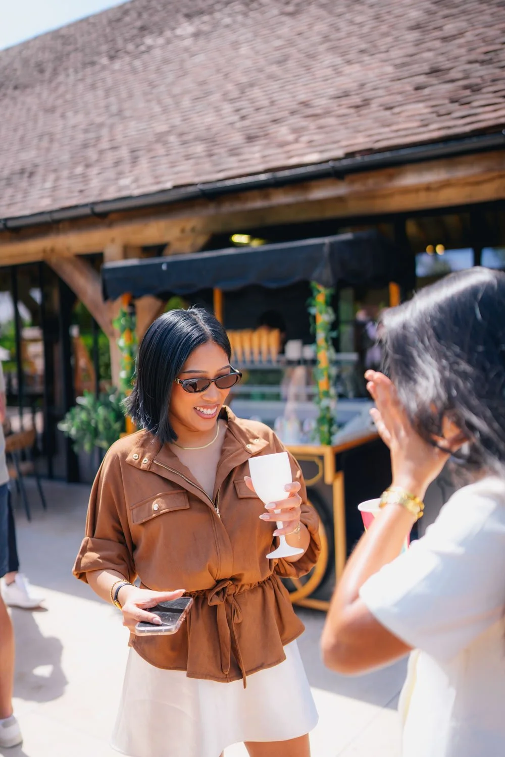 Two women talking and holding drinks at an outdoor event or festival, with a food stall in the background.