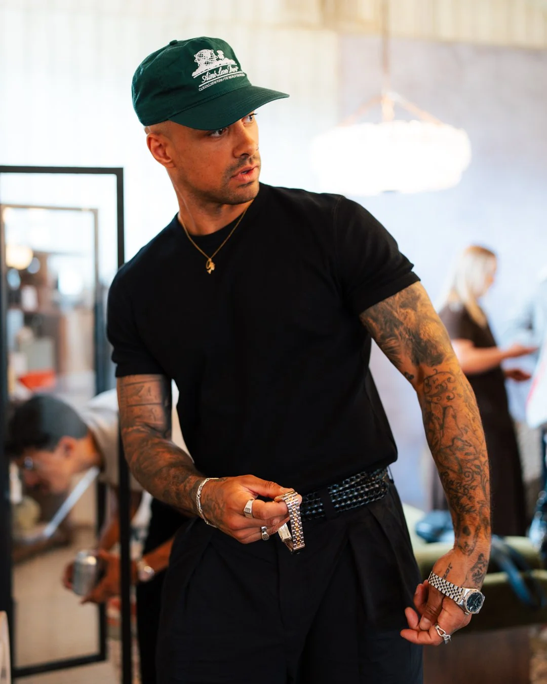 A man wearing a green cap, black t-shirt, and black pants with a diamond-studded belt is standing indoors with his left arm bent, showing multiple tattoos, rings, a watch, and a bracelet. There are people in the background, including a woman wearing 