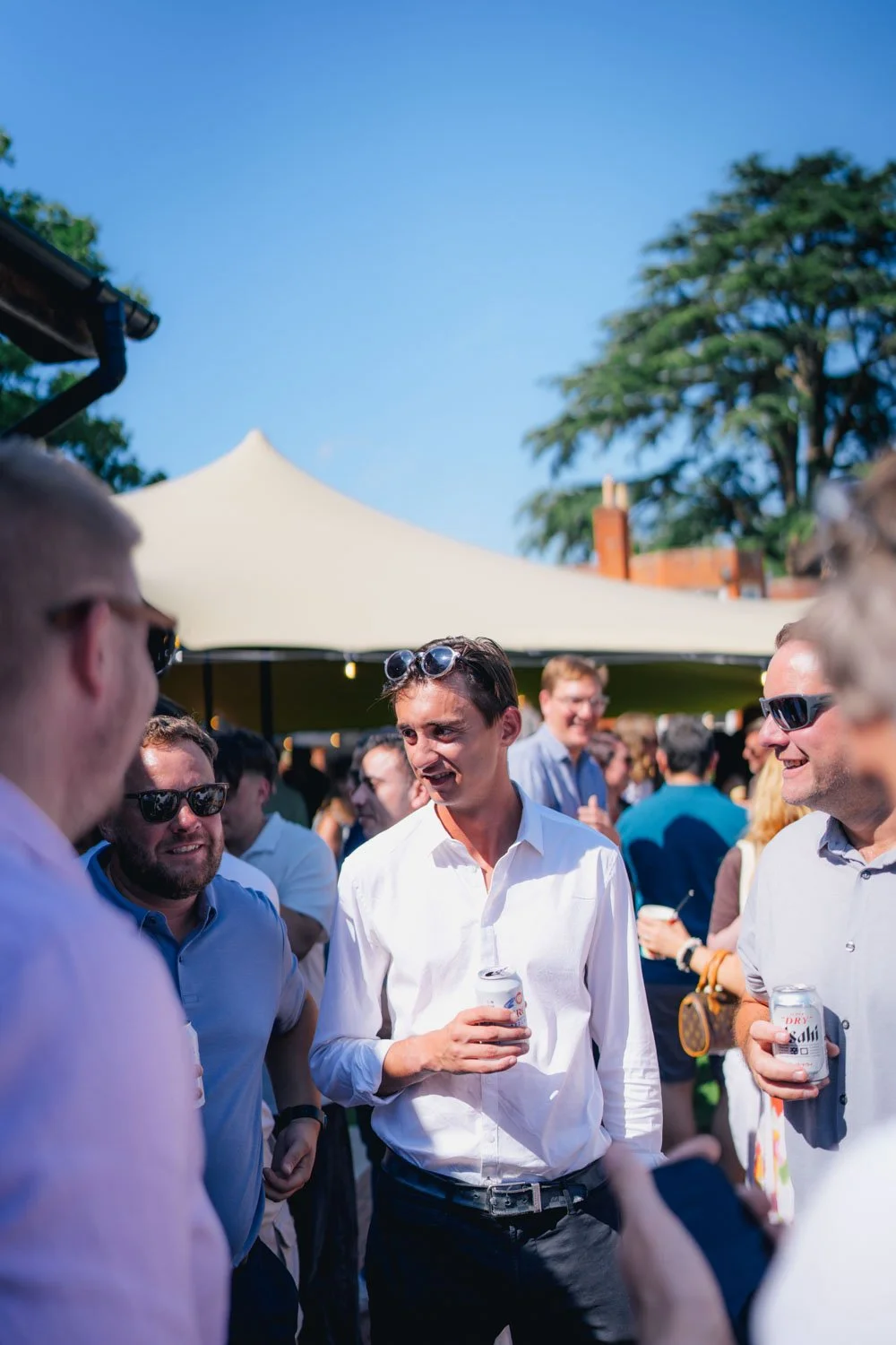 Group of men at outdoor social event, some wearing sunglasses, holding drinks, engaging in conversation, with a large canopy tent and trees in the background on a sunny day.