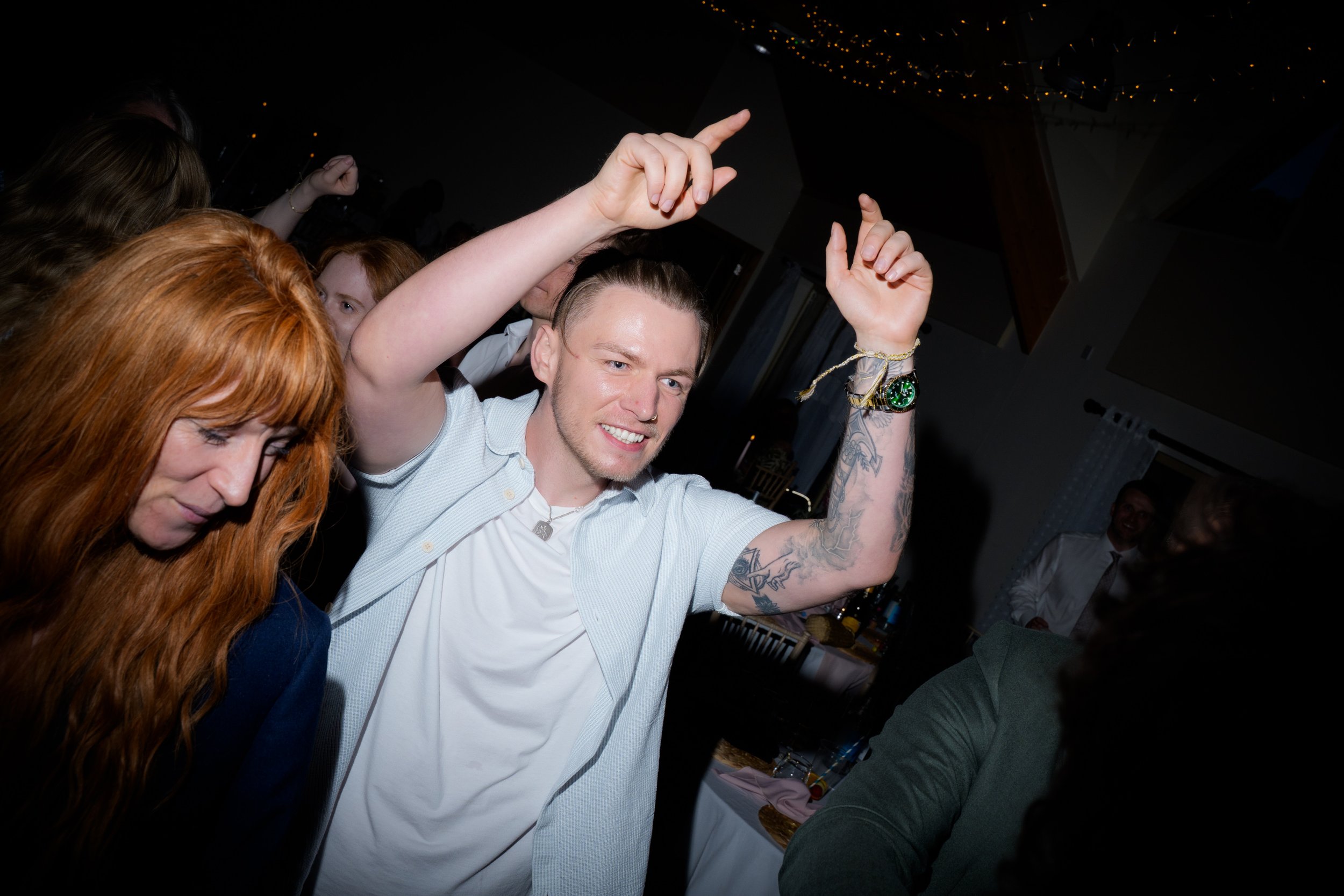 A young man with tattoos on his arms dancing with others at a party, raising his right arm, with people around him, and string lights hanging overhead.