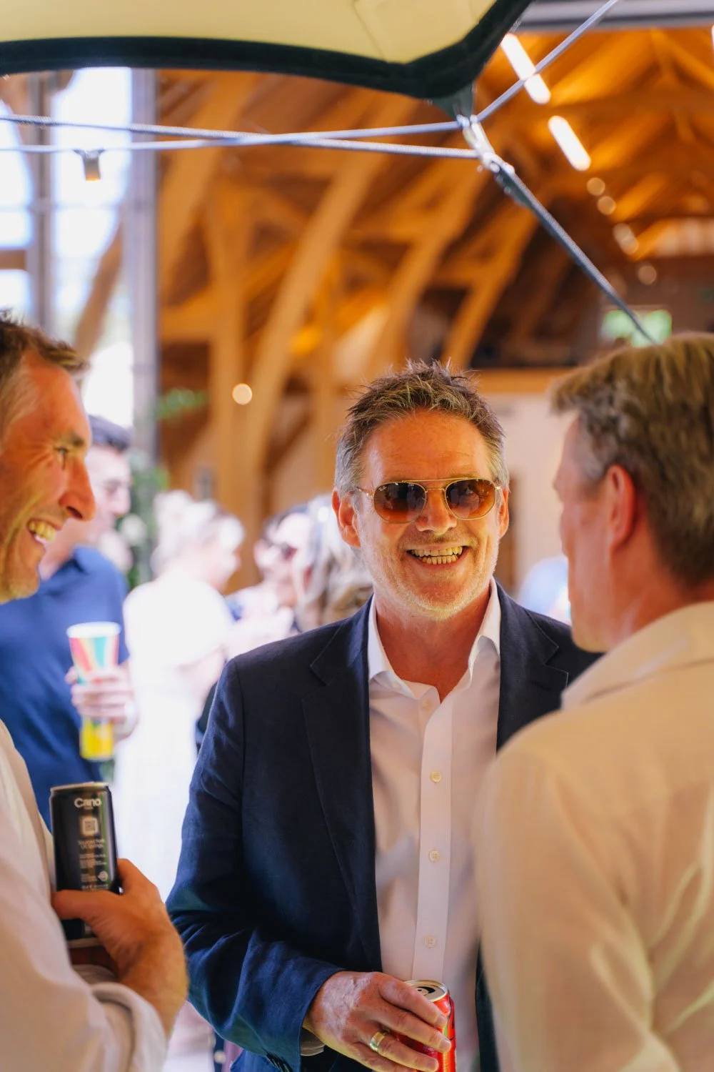 Group of people socializing at a party or event, with three men in the foreground engaged in conversation, smiling, holding cans of drinks, inside a venue with wooden beams and warm lighting.