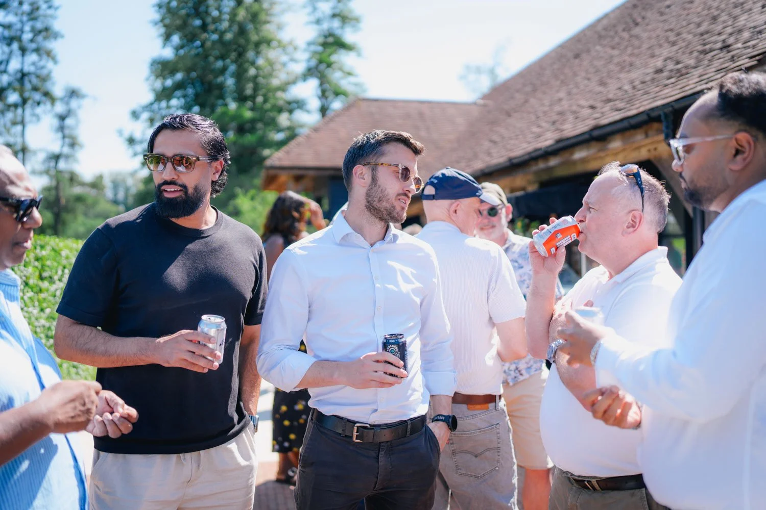 Group of men socializing outdoors, some drinking soda, at a sunny gathering near a wooden building with trees in the background.